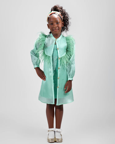 Girl in mint glitter jersey dress with silk organza overshirt and feather trim.