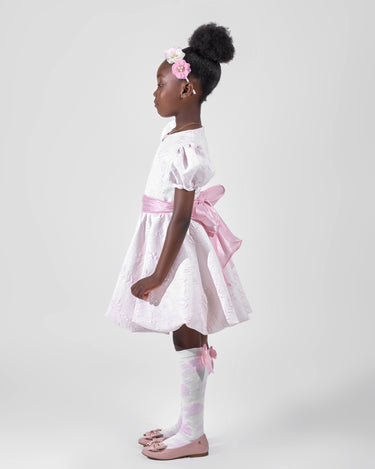 Girl in blush pink floral brocade dress with puff sleeves and organza bow.